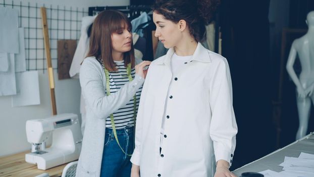 Pretty customer is trying on tailored shirt and sharing her opinion while female clothing designer is measuring and checking new garment and taking to client in light studio.