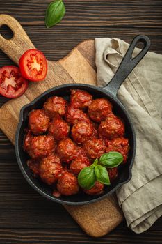 Top view of delicious meatballs with tomato sauce and fresh basil in cast iron rustic vintage pan served on cutting board, wooden background. Tasty homemade meatballs