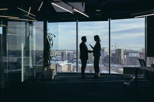 Silhouettes of two employees team of businessmen, Asian man and woman are consulting and communicating, working in a modern office by the window