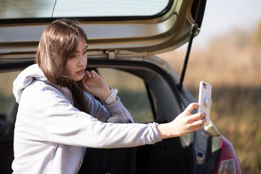 A beautiful girl sits in a car and looks at the phone screen. Taking a selfie in nature or streaming.