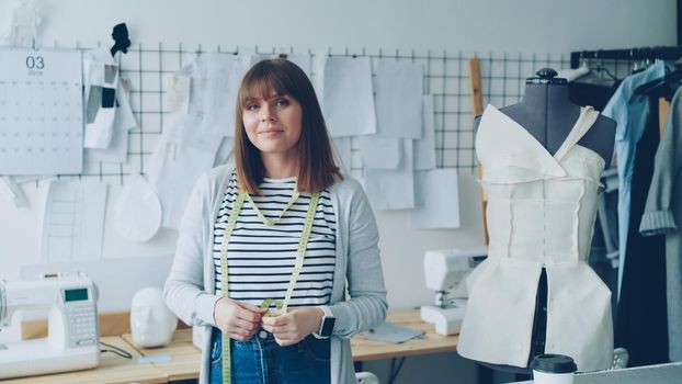 Portrait of beautiful young woman seamstress standing in workplace, touching measuring tape and looking at camera. Dummy, women's garments, sketches and sewing machine are visible.