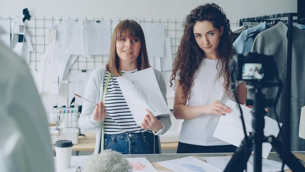 Creative bloggers tailors recording video about new sketches. Geirls are greeting followers, showing drawings and talking emotionally. Camera, garments and tailoring items are visible.