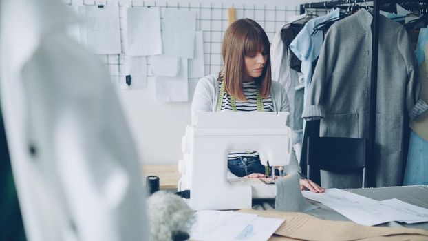 Self-employed female clothing designer is working with sewing machine, stitching textile and looking at sketches lying on studio table. Everyday work at tailor's shop concept.