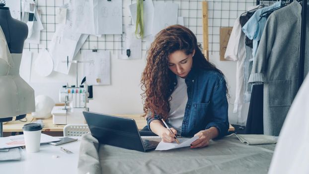 Self-employed seamstress is working with laptop sitting at table in modern light workshop. Woman is drawing garment sketch. Browsing for inspiration concept.