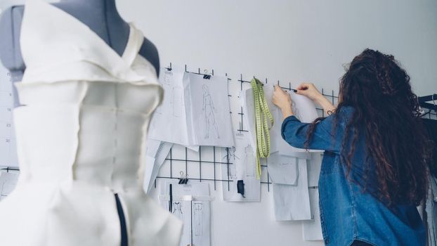Young female clothing designer is looking at sketches and hanging drawings on wall in light workshop. Large collection of drafts above tailoring desk, clothed dummy in foreground.