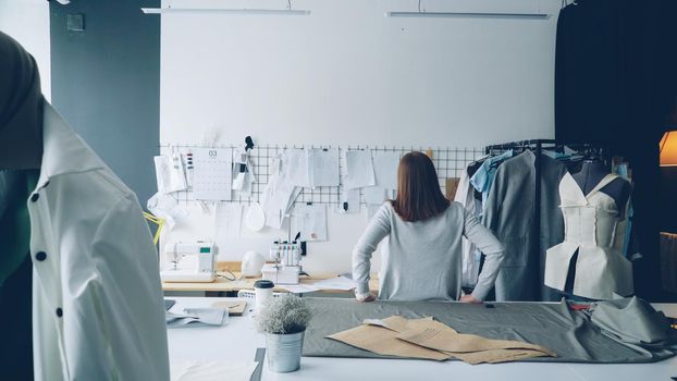 Young female designer standing with her back to camera and looking at drawings hanging on white wall. Attractive woman is thinking about new clothing collection.