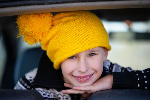 Positive little boy in a yellow knitted hat looks at the camera and smiles. Portrait of a ten year old boy. Child in the car window.