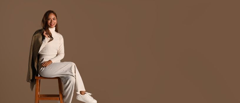 Panoramic shot of stylish woman holding trench coat sitting on wooden chair isolated on brown background. Autumn fashion concept.