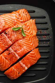 Top view, close-up of cut in slices fresh raw salmon fillet garnished with green basil leaves on grill skillet, gray stone background. Preparing salmon fillet for cooking, healthy eating concept