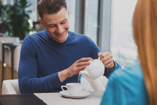 Cheerful handsome man smiling joyfully, pouring tea into his cup, having breakfast with his girlfriend at the local restaurant. Lovely couple enjoying morning tea at the cafe. Love, coziness concept