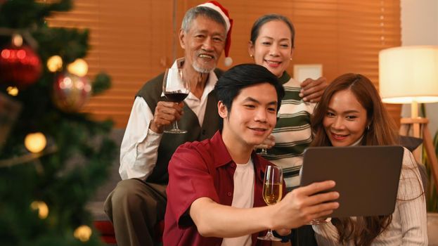 Young couple using digital tablet, taking a selfie with grandparents in living room decorated for celebrating New year and Christmas festive.