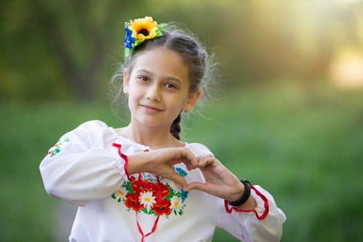 A little Ukrainian and Belarusian girl in an embroidered shirt shows a heart sign with her fingers.