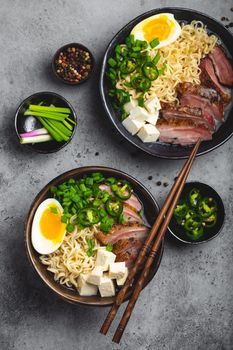 Two bowls of tasty Asian noodle soup ramen with meat broth, tofu, pork, egg with yolk on grey rustic concrete background, close up, top view. Hot tasty Japanese ramen soup for dinner asian style