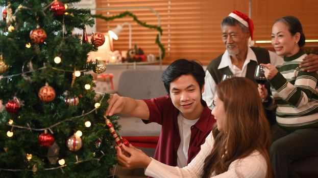 Loving young couple decorating Christmas tree, preparing for celebrating winter holidays together in cozy living room.