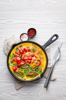 Healthy frittata in cast iron pan with fried beaten eggs and seasonal vegetables on white rustic wooden background. Italian omelette with organic spinach, bell pepper, tomatoes, from above