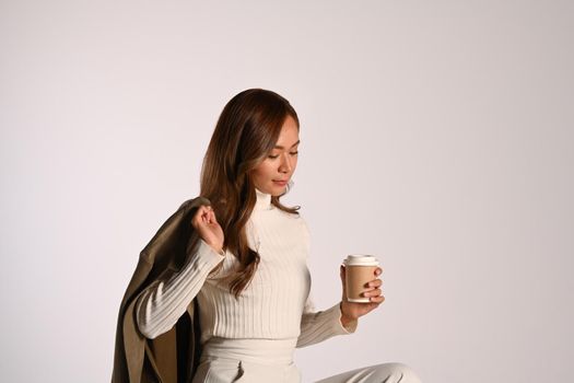 Young beautiful lady in fashionable autumn outfit holding paper cup sitting over light background. Autumn and Winter concept.