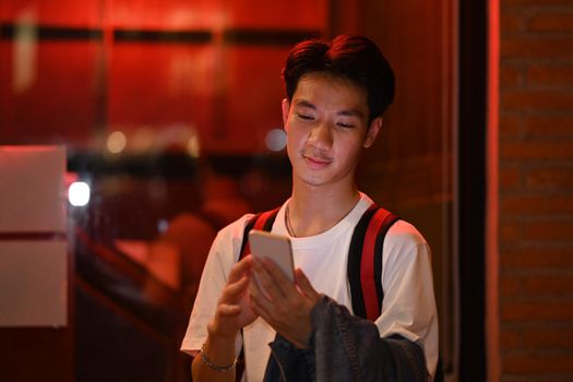 Asian male traveler standing on night city street near showcase and using mobile phone. Night life concept.