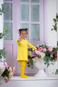 A little African girl in a yellow dress stands on the porch of a house with flowers.