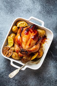 Close-up of baked or roasted whole juicy crispy organic chicken with potatoes, garlic, lemon in white casserole dish with serving spoon on concrete rustic background, top view