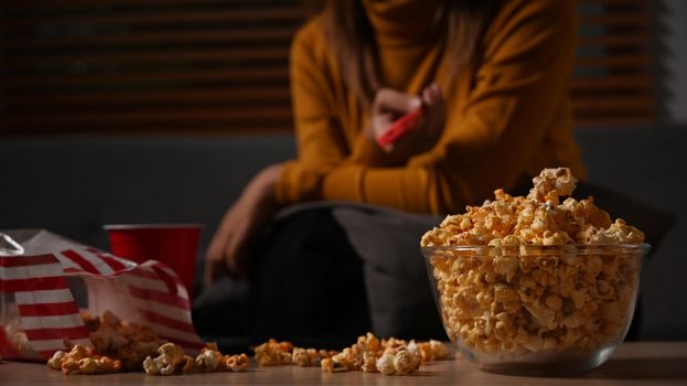 Bowl of popcorn and cup on wooden table with young woman sitting in background. Entertainment and leisure activity concept.