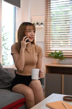 Pretty young woman in casual clothes drinking coffee and talking on mobile phone at cozy home.