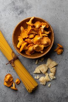 Wild forest mushrooms chanterelles in wooden bowl, raw pasta spaghetti, cheese parmesan on concrete rustic background top view. Seasonal autumn food, ingredients for cooking fresh chanterelles pasta