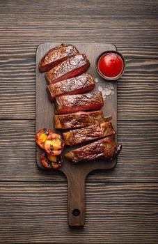 Grilled or fried and sliced marbled meat steak, tomatoes as a side dish and sauce on wooden cutting board, top view, close-up, wooden rustic background. Beef meat steak sirloin concept