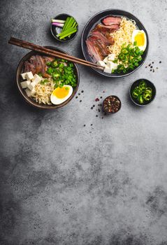 Two bowls of tasty Asian noodle soup ramen with broth, tofu, pork, egg on grey rustic concrete background, space for text, close up, top view. Hot tasty Japanese ramen soup for dinner with copy space