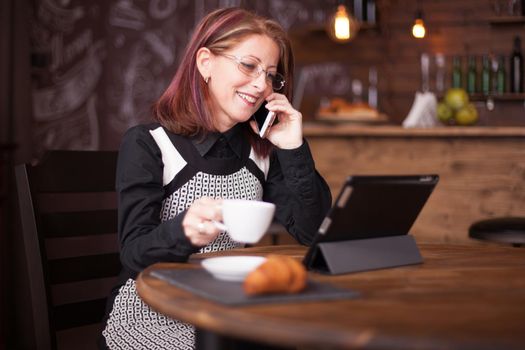 Beautiful smiling adult businesswoman while talking on the phone in front of tablet. Holding a cup of coffe in vintage restaurant