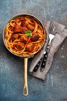 Overhead of traditional homemade pasta with roasted meatballs, tomato sauce in copper vintage pan with fork and knife on rustic concrete background. Classic homemade meatballs spaghetti for dinner