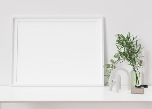 Empty horizontal picture frame standing on white shelf. Frame mock up. Copy space for picture, poster. Template for your artwork. Close up view. Plant in vase, home accessories, sculpture. 3D render