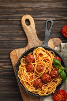 Top view of delicious pasta with meatballs, tomato sauce and fresh basil in cast iron rustic vintage pan served on cutting board, wooden background. Tasty homemade meatballs spaghetti