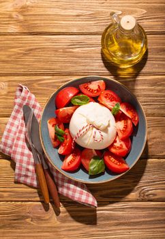 Italian salad with fresh tomatoes, burrata cheese, basil served on ceramic plate with bottle of olive oil on rustic wooden background from above, healthy snack or lunch