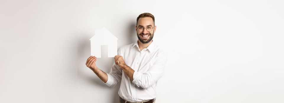 Real estate. Handsome man showing house model and smiling, broker showing apartments, standing over white background.