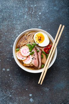 Tasty Japanese noodle soup ramen in white ceramic bowl with meat broth, sliced pork, narutomaki, egg with yolk on rustic stone background. Traditional dish of Japan, top view, close-up, concept