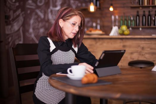 Beautiful smiling adult woman enjoying coffe while working on her tablet. Cozy vintage restaurant