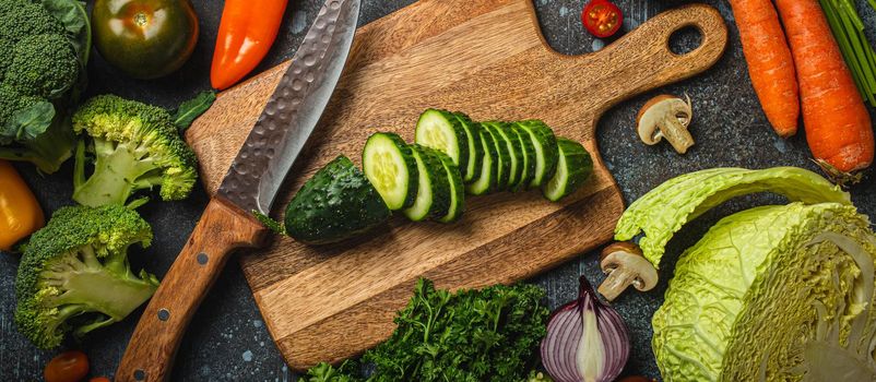 Chopped cucumber on wooden cutting board with knife and assorted fresh vegetables on rustic concrete table, diet and vegetarian food preparing and meal cooking concept, healthy lifestyle