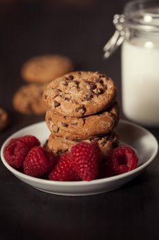 Chocolate chip cookies on white plate dark old wooden table with red raspberry and milk. Delicious breakfast.