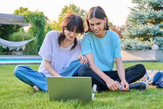 Two teenage friends of students sitting on grass with laptop, in backyard, guy and girl 18 years old study together. Friendship, youth, technology, high school, college, lifestyle concept