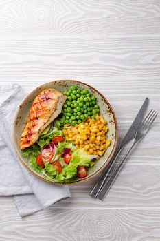 Baked chicken breast on plate with fresh salad, green peas and corn, white wooden background, top view. Healthy fitness meal with chicken fillet, balanced in proteins and carbs. Dieting and nutrition