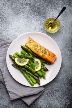 Grilled salmon fillet with green asparagus and seasonings on plate, gray concrete background. Healthy balanced dinner with salmon and asparagus good for diet and wellness, top view, close-up