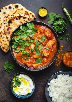 Traditional Indian dish Chicken tikka masala with spicy curry meat in bowl, basmati rice, bread naan, yoghurt raita sauce on rustic dark background, top view, close up. Indian style dinner from above