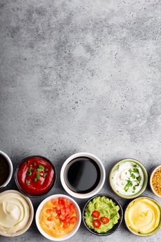 Set of different healthy sauces in bowls on rustic concrete background, top view, close-up. Space for text. Tomato ketchup, mayonnaise, guacamole, mustard, pesto, cheese sauce - assortment of dips