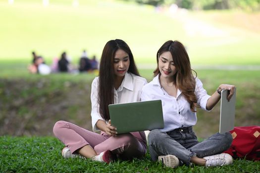 Two young women workers sitting on green grass in the nature park and working on laptop together.