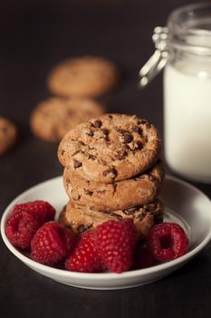 Chocolate chip cookies on white plate dark old wooden table with red raspberry and milk. Delicious breakfast.