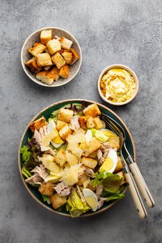 Fresh Caesar salad with lettuce salad, chicken breast, boiled eggs and croutons in ceramic bowl with dressing on the side on gray table. Top view, healthy salad great for lunch, snack or appetizer