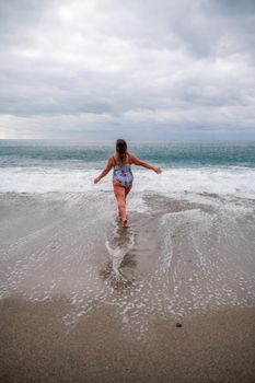 A plump woman in a bathing suit enters the water during the surf. Alone on the beach, Gray sky in the clouds, swimming in winter