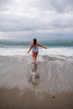 A plump woman in a bathing suit enters the water during the surf. Alone on the beach, Gray sky in the clouds, swimming in winter