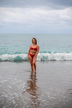 A middle-aged woman with a good figure in a red swimsuit on a pebble beach, running along the shore in the foam of the waves.