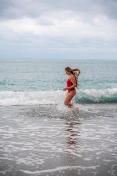 A beautiful and sexy brunette in a red swimsuit on a pebble beach, Running along the shore in the foam of the waves.